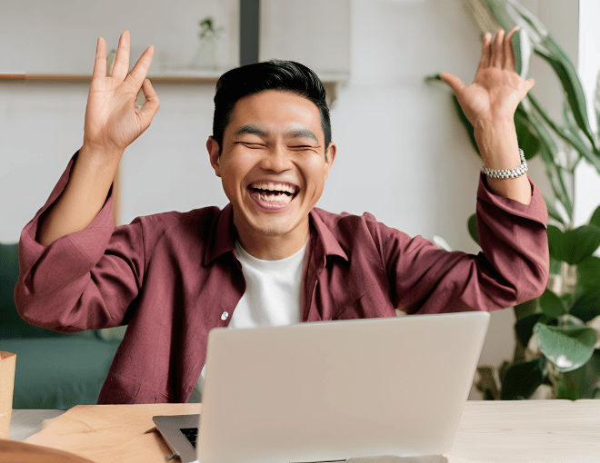 An Indonesian man smiles joyfully at his laptop screen, hands raised in excitement.